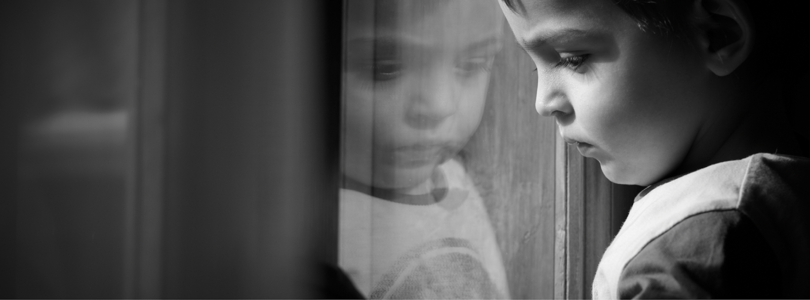 Black-and-white photograph of a child looking down beside a window, with the child's reflection in the glass.